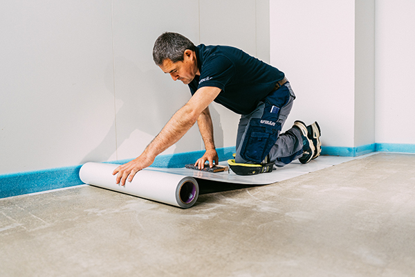 Ein Mann kniet auf dem Boden und rollt eine große weiße Rolle Trockenklebstoff aus der Wand ab, um die Oberfläche für Installations- oder Renovierungsarbeiten vorzubereiten. Er trägt Arbeitskleidung und Knieschützer.