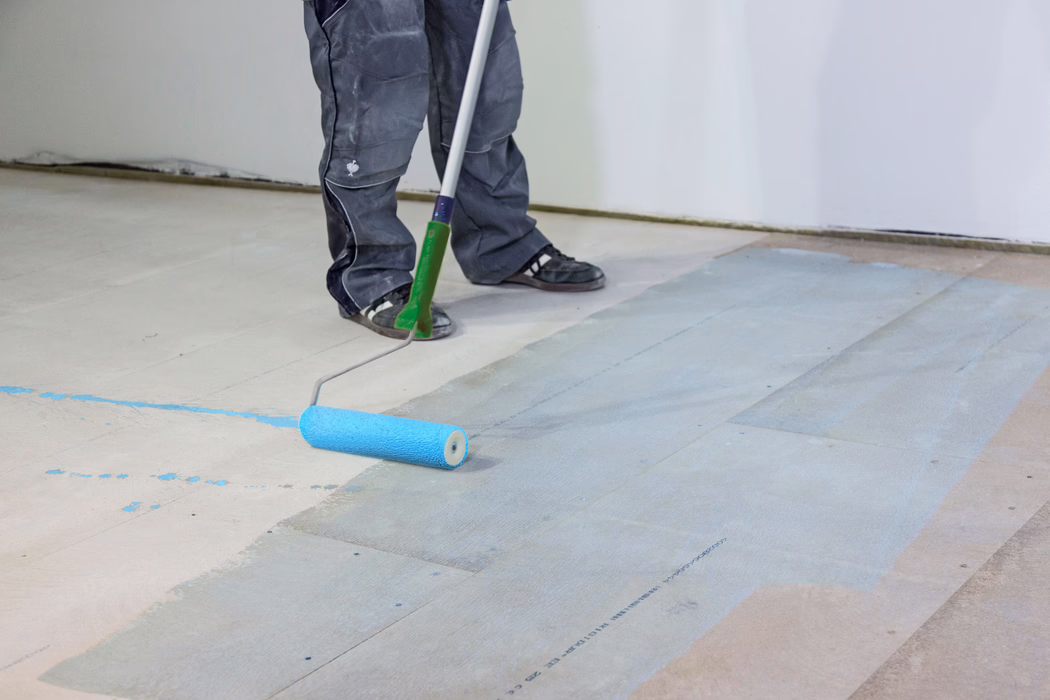 UZIN_Application Image_PE360 Plus_Renoplan System_2020-10 Die Unterschenkel und Füße eines Handwerkers sind zu sehen, während er mit einer langstieligen Farbrolle einen hellblauen Grundanstrich auf einem Holzunterboden aufträgt. Die Person trägt dunkle Arbeitshosen und schwarze Turnschuhe; die Rolle hinterlässt einen feuchten blauen Film auf den Dielen in einem Innenraum mit weißen Wänden. Das Bild konzentriert sich auf die Bodenvorbereitung, Werkzeuge und Farbspritzer.