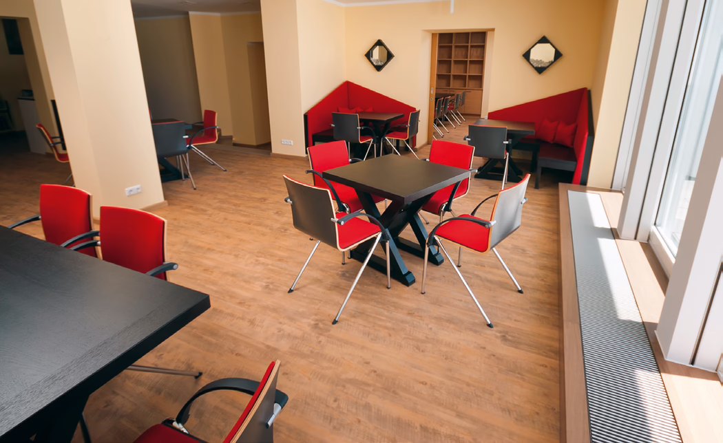 Ein heller, moderner Lounge- oder Essbereich mit hellem Holzfußboden, beigen Wänden, schwarzen quadratischen Tischen und roten gepolsterten Stühlen mit verchromten Beinen. Große Fenster auf der rechten Seite lassen Tageslicht einfallen; eingebaute rote Eckbänke, Regale im Hintergrund und eine Heizkörperabdeckung entlang des Fensters vervollständigen die ordentliche Anordnung, die sich für ungezwungene Treffen oder Mahlzeiten eignet.