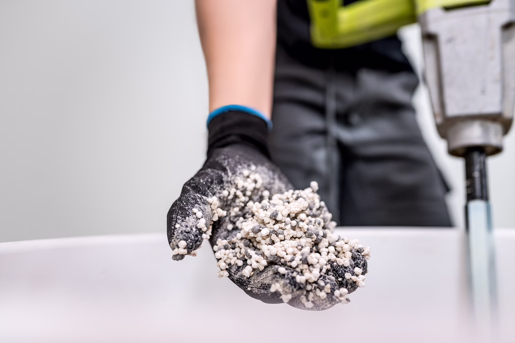 Nahaufnahme einer schwarz-behandschuhten Hand, die eine Handvoll kleiner weißer und grauer Körner hält (z. B. Dünger oder Perlit). Der Handschuh hat einen blauen Saum; im unscharfen Hintergrund ist eine Person in dunkler Kleidung mit gelber Warnweste und ein Bohrgerät zu sehen. Neutrale Innenaufnahme, Fokus auf die Struktur der Granulate und die Hand.