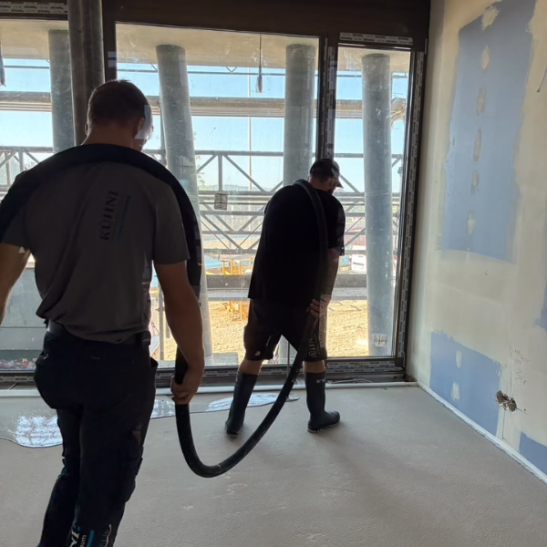 Two workers clean a construction site room with large windows, using a hose to remove debris or water from the floor. Scaffolding and construction materials are visible outside the windows.