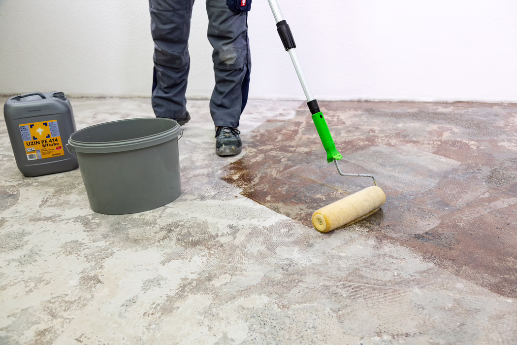 Die unteren Beine und Füße eines Handwerkers sind zu sehen, während er mit einer langstieligen Farbrolle eine braune Grundierung beziehungsweise Klebeschicht auf einen nackten Betonboden aufträgt. Ein grauer Kunststoffeimer und ein grauer Kanister mit der Aufschrift "UZIN PE 414 Biturbo" stehen in der Nähe an einer weißen Wand. Der Handwerker trägt graue Arbeitshosen und schwarze Sicherheitsschuhe; der Boden zeigt verbliebene Kleberreste und geschliffenen Beton, während die Rolle die Flüssigkeit gleichmäßig verteilt, um den Untergrund vorzubereiten.