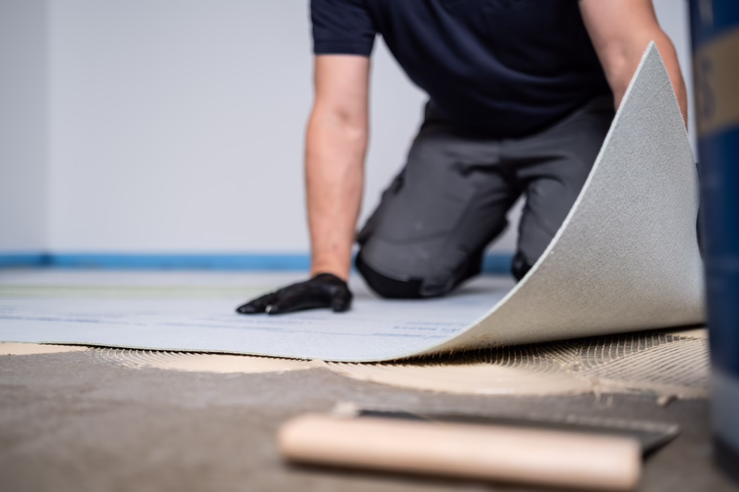 Nahaufnahme eines Handwerkers, der kniend einen Bodenbelag verlegt. Die Person trägt schwarze Handschuhe und Arbeitshosen und hebt eine Unterlagsbahn über frisch aufgetragenen Kleber mit sichtbaren Spuren einer gezahnten Kelle; geringe Schärfentiefe zeigt ein modernes Innenraum und Werkzeuge in der Nähe.