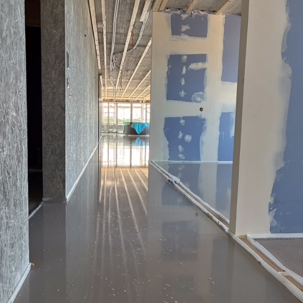 A hallway in a building under construction with unfinished walls, exposed ceiling, and a shiny, freshly poured floor reflecting the light from large windows at the end.