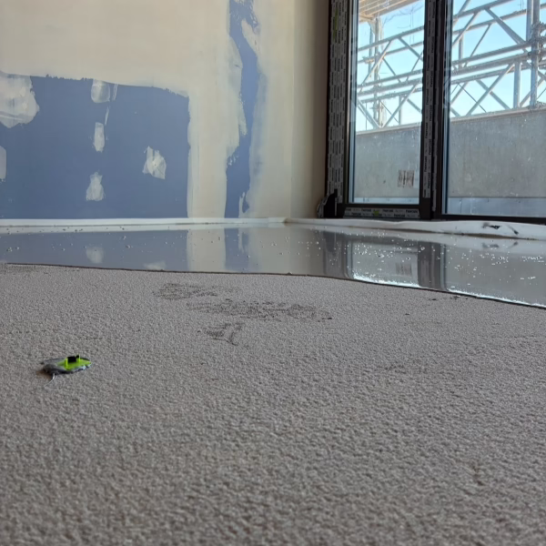 A flooded room with water pooling near large windows, partially on carpet. The carpet is wet and stained, with a small green and silver item lying on it. Walls show patches of unfinished paint.