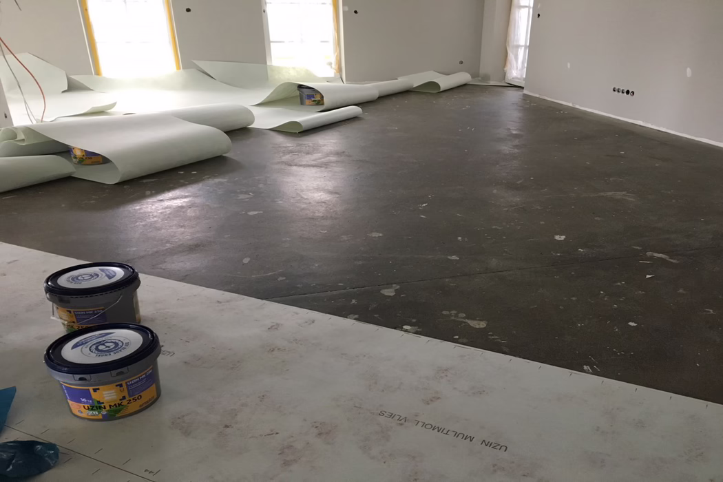 UZIN_Application_Image_Multimoll Vlies_2019-06 Innenraum einer Wohnung in Renovierung: großer offener Raum mit dunklem, noch nicht fertigem Betonunterboden, übersät mit Farbspritzern, Fußabdrücken und Ausbesserungen. An der Rückwand liegen mehrere breite Rollen hellgrüner Unterlage oder Bodenbahn teilweise ausgerollt bei mit Folie geschützten Fenstern. Im Vordergrund stehen zwei Kunststoffeimer mit Klebstoff oder Grundierung (gekennzeichnet UZIN MK 250) auf Schutzplatten; die Wände sind frisch verputzt oder grundiert in hellgrau und zeigen offene Elektroanschlüsse und Steckdosen. Diffuses Tageslicht fällt durch die abgedeckten Fenster und erzeugt weiche Spiegelungen auf dem Boden; keine Arbeiter oder Werkzeuge sind sichtbar, was auf eine unterbrochene Installationsphase hindeutet.