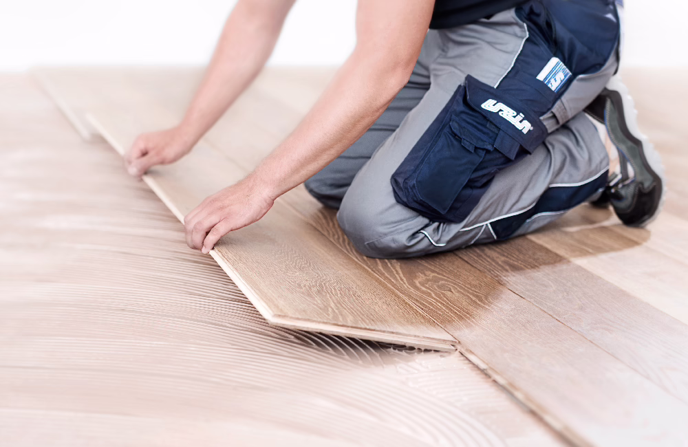UZIN_Application Image_Parquet_2016-01 Ein Handwerker kniet auf dem Boden und verlegt helle Eichen-Laminatdielen; Nahaufnahme der Hände, die eine Diele über Kleber anpassen, er trägt graue und marineblaue Arbeitshosen mit Taschen und Knieschonern; Innenraum, gleichmäßige Beleuchtung, versetztes Dielenmuster.