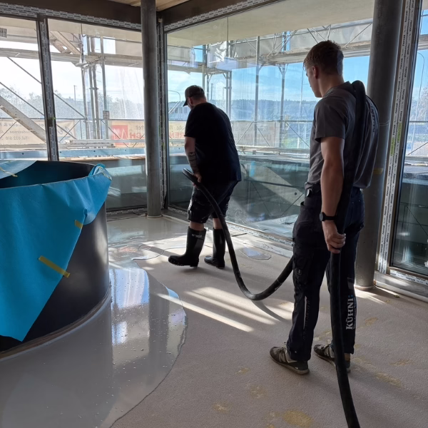 Two workers in a sunlit room use hoses to spread liquid flooring material. One wears boots and operates the hose, while the other stands nearby holding a second hose. A large covered object is beside them.