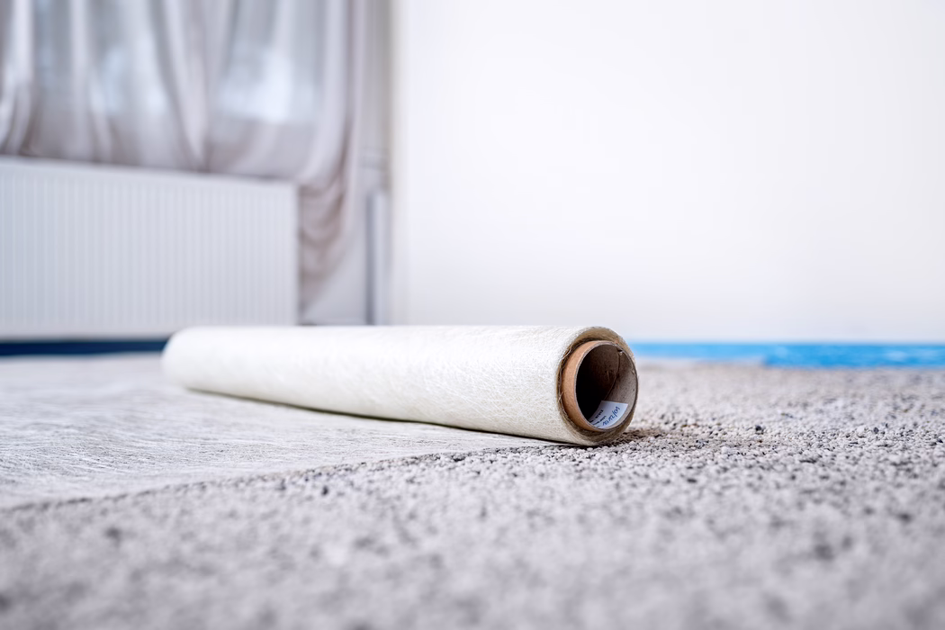 Nahaufnahme einer teilweise abgerollten Tapete oder Bodenunterlage, die auf einem hellen Teppich in einem hellen, spärlich eingerichteten Raum liegt. Der Fokus liegt auf dem Pappkern des Rollenendes im Vordergrund, während im unscharfen Hintergrund ein Heizkörper und transparente Vorhänge als weiches Bokeh zu sehen sind. Neutrale Farbtöne und natürliches Licht schaffen eine ruhige, minimalistische Atmosphäre und deuten auf Renovierungsarbeiten oder eine laufende Innenraumgestaltung hin.