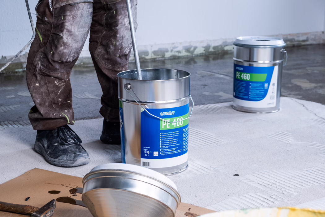 Die Unterschenkel und Füße eines Bauarbeiters stehen auf einem teils vorbereiteten Betonboden neben geöffneten Metallbehältern mit Bodenkleber, beschriftet mit 'PE 460'. Ein Eimer wird mit einer langen Rührstange umgerührt, Deckel und Werkzeug liegen in der Nähe auf dem Boden. Der Arbeiter trägt abgenutzte, mit Farbe und Staub besprenkelte Arbeitshosen und lädierte Arbeitsschuhe. Die Betonschicht zeigt ein strukturiertes Klebermuster und unebene Kanten an der Wand, was auf laufende Bodenarbeiten in einem unfertigen Raum mit neutraler Beleuchtung hinweist.