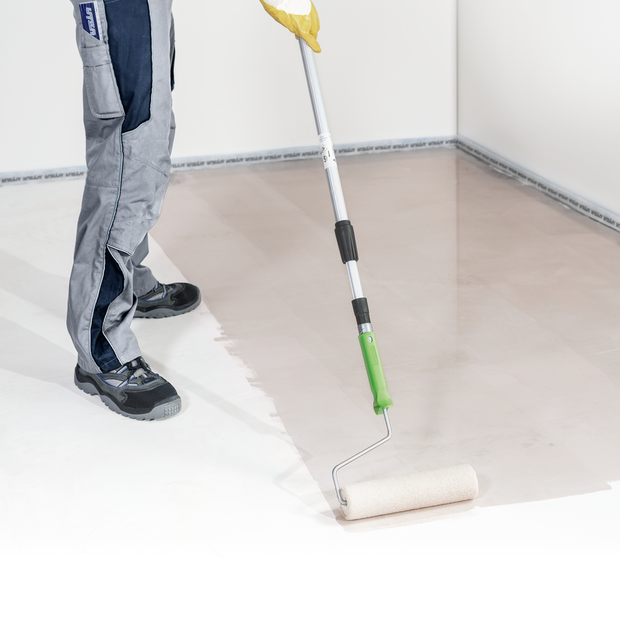 Eine Person mit Arbeitshandschuhen und Schutzkleidung trägt mit einer Farbrolle an einer ausziehbaren Stange eine Beschichtung auf den Boden eines teilweise fertiggestellten, weißwandigen Raums auf.