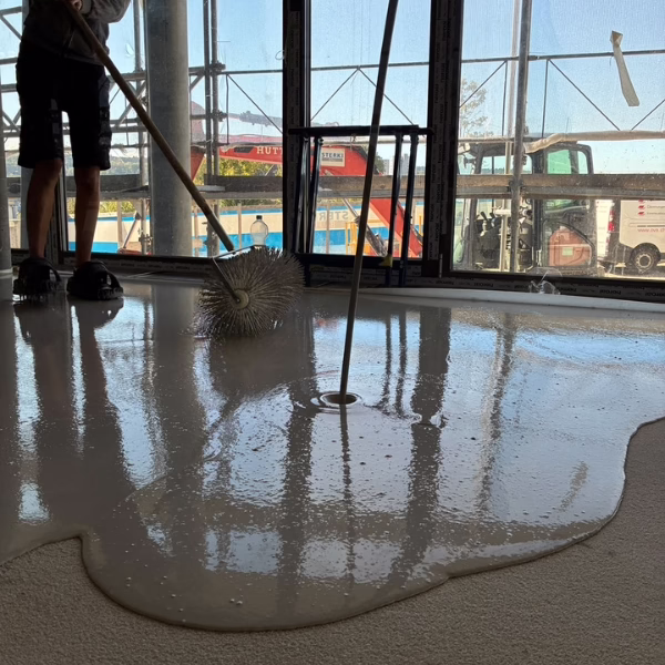 A person uses a spiked roller to spread and smooth self-leveling floor compound on a construction site, with scaffolding and vehicles visible outside large windows.