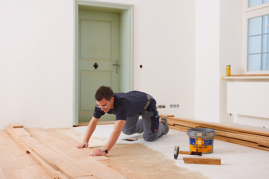 Ein Handwerker mit Schutzbrille und Arbeitskleidung kniet auf einem teilweise verlegten Holzboden und drückt eine Diele in den Kleber; Werkzeuge und Materialien wie ein Klebereimer, ein Hammer und gestapelte Bodenbretter liegen in der Nähe in einem hellen, unmöblierten Raum mit blassen Wänden und einer geschlossenen Tür.