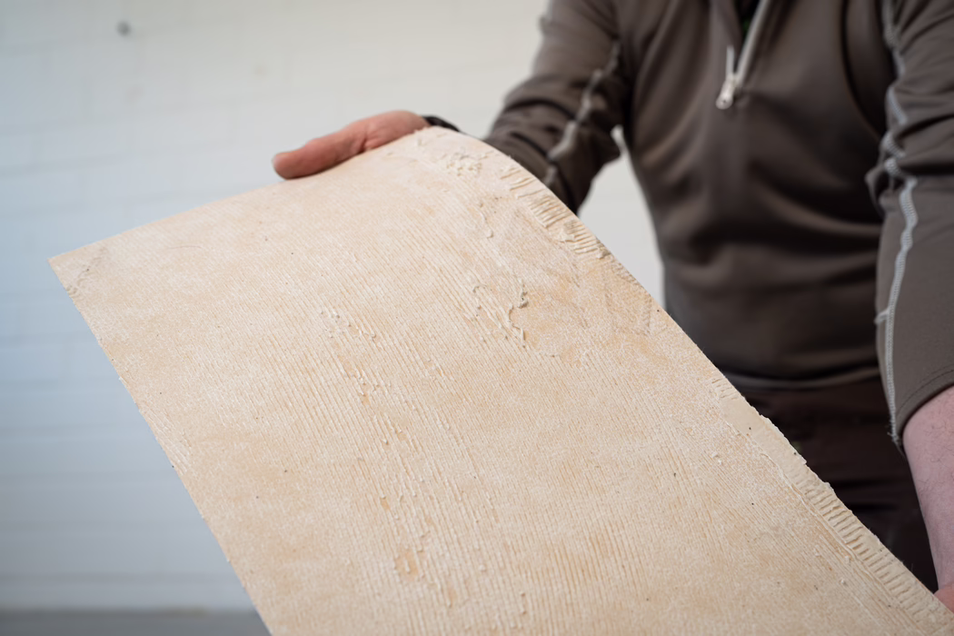 Nahaufnahme einer Person, die ein großes rechteckiges, beigefarbenes Brett mit strukturierten Klebespuren hält; Rumpf und Hände sind teilweise sichtbar, die Person trägt ein braunes Langarmshirt. Auf dem Brett sind gerillte Klebelinien und kleine trockene Putz- oder Klebereste zu erkennen.