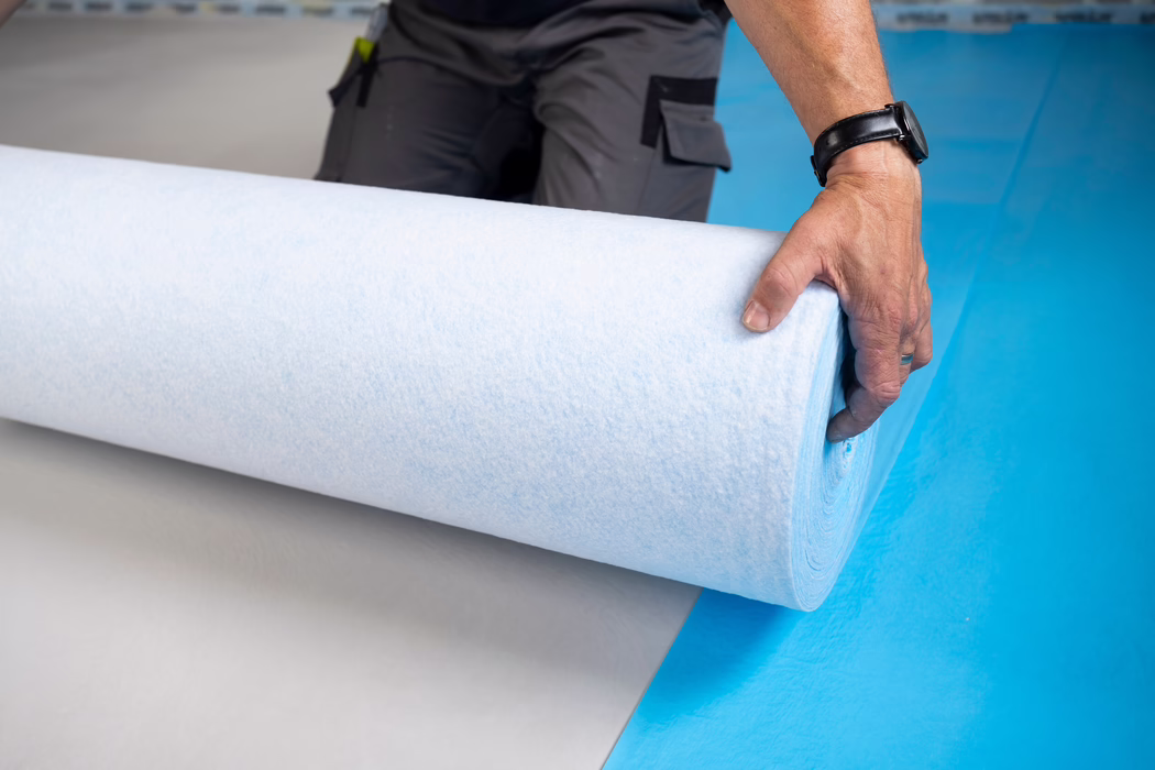Nahaufnahme der Hand eines Arbeiters, der eine große hellblau-weiße Unterlage- bzw. Isolationsrolle auf einem kräftig blauen Boden abrollt. Die rechte Hand trägt eine schwarze Armbanduhr und einen schlichten Ring und hält den Rand der Rolle; graue Arbeitshosen mit Taschen und ein eingestecktes Werkzeug sind im Hintergrund teilweise sichtbar. Die weiche, faserige Oberfläche des Materials ist erkennbar, die Szene deutet auf eine Innenraum-Baustelle oder Bodenverlegung hin und die Komposition betont die Rundung der Rolle sowie den Farbkontrast zwischen Weiß, hellblau und dem kräftigen Blau des Bodens.