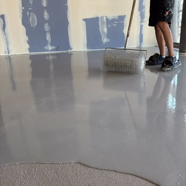 Person using a spiked roller to spread and level gray epoxy floor coating in a room with unfinished drywall.