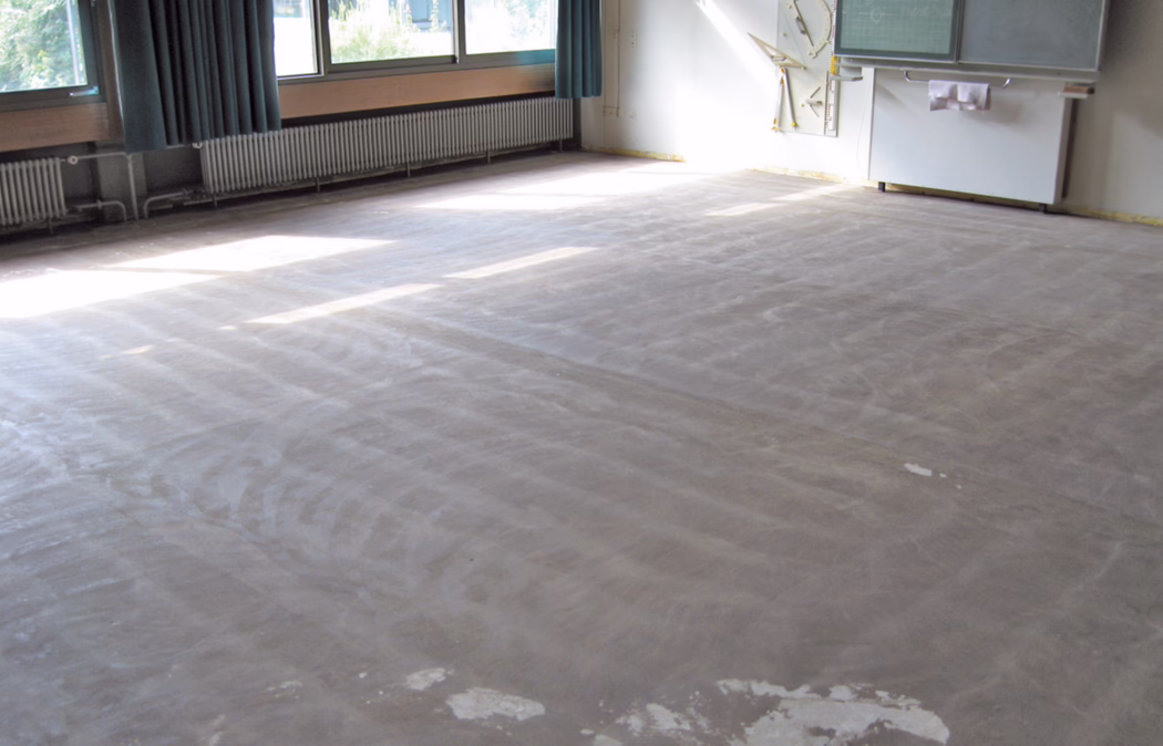 Ein helles, leeres Klassenzimmer, in dem der Bodenbelag entfernt wurde und nun ein staubiger, roher Unterboden sichtbar ist. Große Fenster an der linken Seite lassen Licht herein, das rechteckige Lichtflecken auf dem Boden erzeugt; blau-grüne Vorhänge sind teilweise zugezogen, Heizkörper verlaufen unter den Fenstern. Vorn rechts befinden sich eine Tafel/Whiteboard und montierte Lehrmittel; Reste von Kleber oder Putz sind auf dem Boden zu sehen. Der Raum ist möbelfrei und wirkt, als befände er sich in Renovierung oder intensiver Reinigung.