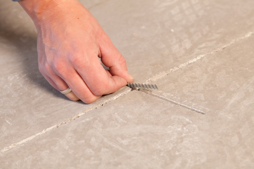 Nahaufnahme der linken Hand einer Person mit einem schlichten Silberring, die einen kleinen gezackten Kunststoff-Fugenkeil aus einer Fuge zwischen hellbeigen bis grauen Bodenplatten zieht. Der Boden zeigt gerade Fugen und eine leicht strukturierte Oberfläche; Hand und Keil sind leicht staubig. Weiches, neutrales Licht betont Hautstruktur und Fliesenoberfläche.
