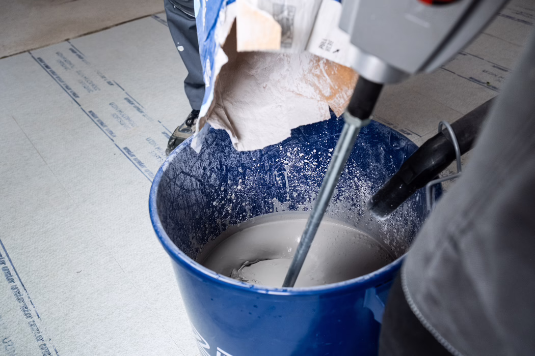 UZIN_Application Image_Levelling Compounds_NC580_2024-03 (22) Nahaufnahme eines blauen Plastik-Eimers auf dem Boden, gefüllt mit grauem Mörtel oder Spachtel, der mit einem elektrischen Rührgerät gemischt wird; ein Papiersack mit Trockenpulver wird eingegossen, Spritzer bedecken die Innenwände, und Beine sowie Werkzeuge eines Arbeiters sind teilweise sichtbar.