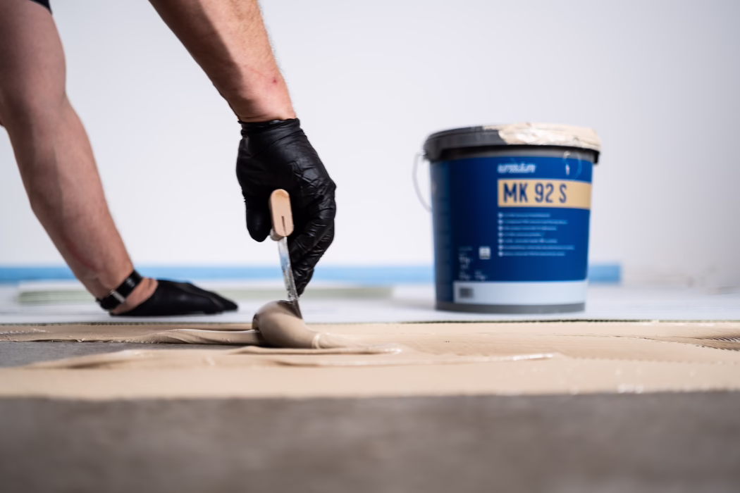 Nahaufnahme einer Person, die mit einer Kelle beigen Bodenkleber oder eine selbstnivellierende Masse auf den Unterboden aufträgt. Die arbeitende Person trägt schwarze Einweghandschuhe; es sind nur Unterarm und Hand in einer Aufnahme aus niedriger Perspektive sichtbar. Im Hintergrund unscharf ein blaues Eimer mit der Aufschrift 'MK 92 S'. Innere Renovierungsszene, Fokus auf die Textur der nassen Masse und die glättende Bewegung der Kelle.