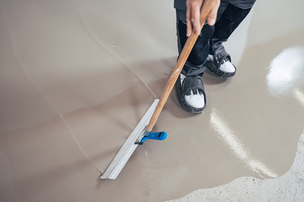 Nahaufnahme eines Arbeiters, der mit einer langstieligen Glättkelle frisch aufgetragenen selbstnivellierenden Estrich glättet; Schutzschuhe sind zu sehen und die Oberfläche wirkt glänzend und nass.