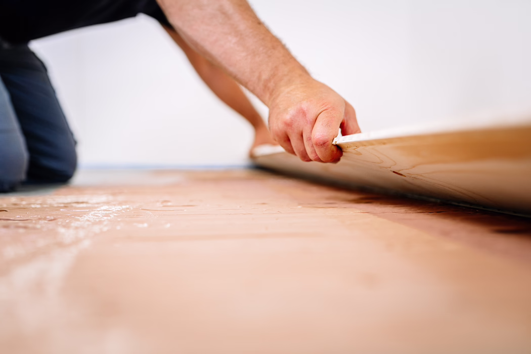 Detailaufnahme einer Person, die Boden verlegt: Die Hand eines knieenden Handwerkers fasst die Kante einer Laminat- oder Holzplanke, während sie auf den vorbereiteten Unterboden gelegt wird. Die geringe Schärfentiefe betont die strukturierte Plankenkante und die Hand, im Hintergrund ist eine weiße Wand und die kniende Haltung erkennbar. Die Oberfläche darunter wirkt wie eine Unterlage oder Klebeschicht mit leicht rosa Tönung; die Szene vermittelt sorgfältige manuelle Arbeit bei einer Renovierung.
