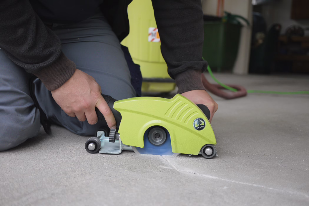 Eine Person kniet auf einem Betonboden und bedient einen kompakten, grünen handgeführten Betonschneider. Die blaue Kreissäge schneidet eine flache Fuge in die Oberfläche, während die Person die Griffe festhält; kleine Führungsräder und eine verstellbare Metallhalterung stützen das Gerät. Im Hintergrund sind ein flexibler Schlauch und Werkstattgeräte zu sehen. Das Foto konzentriert sich auf die Hände und das Schneidwerkzeug; kein Gesicht ist zu sehen.
