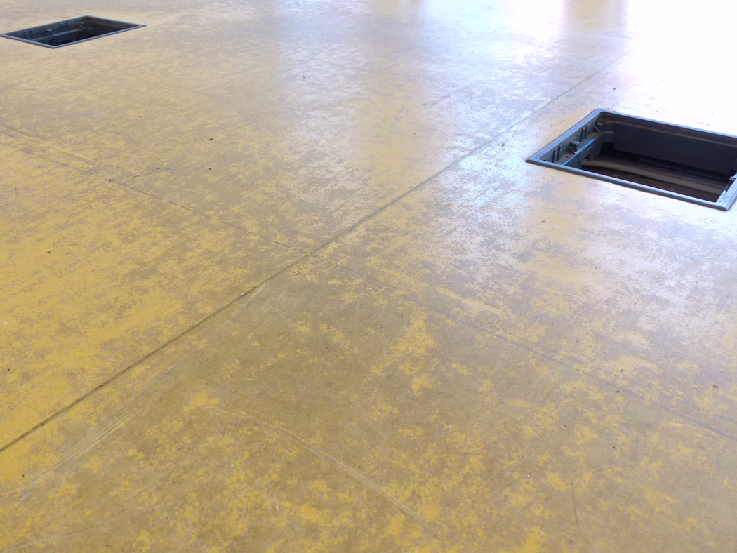 UZIN_Application Image_Basic Cleaner before_2016-09 Ein abgenutzter gelber Linoleum- oder Vinylboden in einem Industrie- oder Büroumfeld, nah aufgenommen. Zwei rechteckige eingelassene Bodendosen (Kabelzugänge) sind erkennbar – eine oben links und eine am rechten Rand mit sichtbaren Kabeln und Kunststoffrahmen. Die Oberfläche zeigt Nähte, Gebrauchsspuren, Schmutzpartikel und eine fleckige Textur; weiches, diffuses Licht spiegelt sich auf dem Boden. Keine Personen im Bild; die Aufnahme betont die Struktur, Abnutzung und die funktionale Nutzung des Bodens.