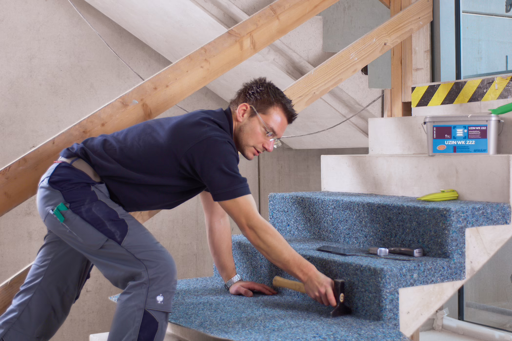 Ein Handwerker verlegt einen blauen, recycelten Teppichbelag auf eine unvollendete Betontreppe in einem renovierungsbedürftigen Innenraum. Er kniet und beugt sich nach vorn, trägt eine Schutzbrille, ein dunkelblaues Polohemd und graue Arbeitshosen und verwendet einen Gummihammer, um den Teppich fest auf den Stufen anzubringen; in der Nähe liegen eine Glättkelle, ein Maßband und ein Plastikeimer mit der Aufschrift ‚UZIN WK 222‘. Die Szene ist hell mit natürlichem Licht ausgeleuchtet, provisorische Holzbretter als Geländerstützen sind sichtbar, der Schwerpunkt liegt auf der sorgfältigen handwerklichen Arbeit und der Textur des Teppichs auf den Stufentritten.