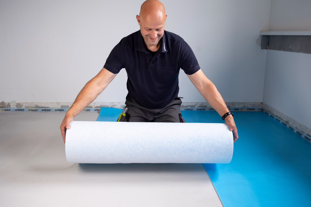 Ein lächelnder Mann mittleren Alters mit kahlem Kopf in einem dunkelblauen Poloshirt rollt eine weiße Unterlagsmatte aus, während er in einem leeren Raum den Boden über einer blauen Dampfbremse verlegt.