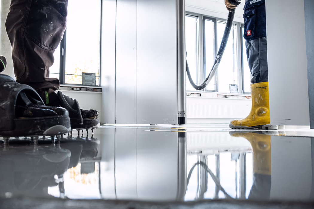 Zwei Bauarbeiter egalisieren einen frisch gegossenen, glänzenden Boden in einem Innenraum. Der linke Arbeiter trägt Nadelsohlen, um auf der nassen Ausgleichsmasse zu laufen; der rechte Arbeiter in gelben Gummistiefeln hält einen Schlauch, der die flüssige Bodenmasse abgibt. Große Fenster und Spiegelungen erzeugen helles Tageslicht; Werkzeuge und ein Heizkörper sind im Hintergrund sichtbar.