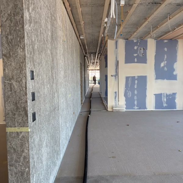 A building interior under construction, with unfinished walls, exposed ceiling beams, visible wiring, and construction materials. A person is seen in the distance through a hallway.
