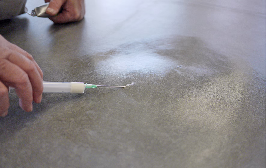 UZIN_Application_Image_RK 36_1_2018-03 Nahaufnahme von zwei Händen, die eine kleine Rissstelle in einer glatten grauen Vinylfläche reparieren; eine Hand hält eine Spritze mit dünner Metallnadel, die in den Riss geführt ist, die andere hält ein kleines Metallwerkzeug oder einen Löffel. Die Oberfläche zeigt eine dezente marmorierte Struktur und weiche Lichtreflexe. Geringe Schärfentiefe richtet den Fokus auf die Nadelspritze und die Rissstelle, während Hintergrund und Hände leicht unscharf sind. Es sind keine Gesichter erkennbar.