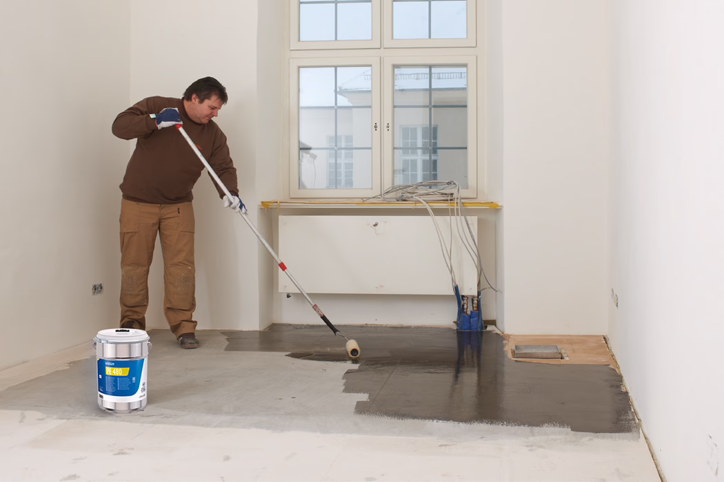 Ein Handwerker in braunem Pullover und beigen Arbeitshosen trägt mit einer langen Rolle eine dunkelgraue Ausgleichsmasse auf den Boden eines leeren Zimmers auf; ein Eimer des Produkts steht auf dem Boden, Kabel liegen auf der Fensterbank, und weiße Wände sowie ein großes Fenster sind sichtbar.