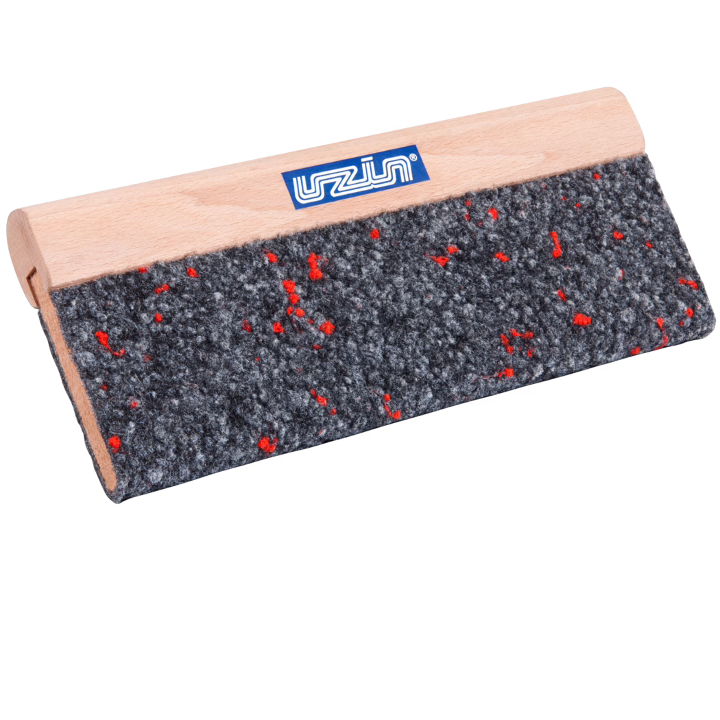 Nahaufnahme eines rechteckigen Werkzeugs mit einem glatten hellen Buchenholzgriff und einer groben grauen Filzauflage, durchsetzt mit kleinen roten Fasern; auf dem Griff ist ein kleines blaues Markenetikett sichtbar. Das Werkzeug ist schräg vor weißem Hintergrund fotografiert, wodurch die Filzstruktur und die feine Holzmaserung betont werden.