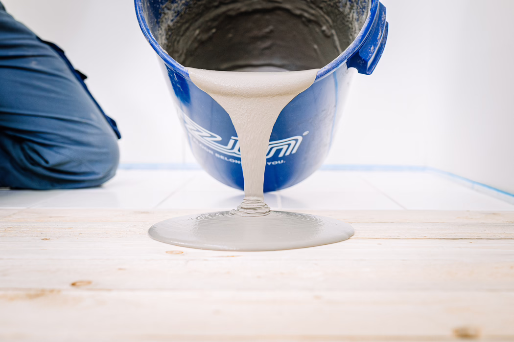 Nahaufnahme eines blauen Eimers, der graue, selbstnivellierende Bodenmasse auf einen Holzunterboden gießt. Die zähflüssige Masse bildet auf dem hellen Holz eine glatte, runde Pfütze; links ist das Knie einer arbeitenden Person in dunkler Arbeitskleidung sichtbar, im Hintergrund eine weiße geflieste Wand und blaues Malerband.