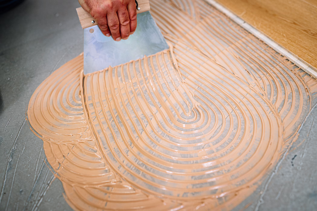 Nahaufnahme einer Hand, die einen gezahnten Fliesenspachtel hält und beige Fliesenkleber in gleichmäßigen parallelen Rillen auf einen grauen Betonboden aufträgt. Die Kleberspuren bilden gebogene, konzentrische Linien; oben am Bildrand ist eine Holzfußbodenleiste sichtbar, die die Vorbereitung zum Verlegen des Bodens zeigt.