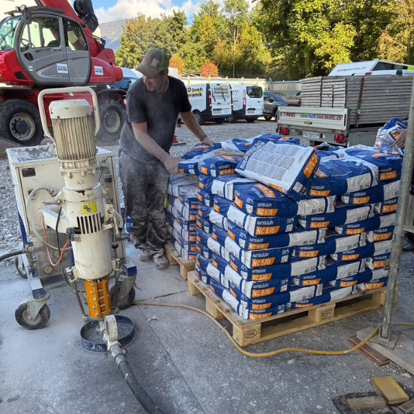 A worker stands next to stacked bags of cement on a pallet at a construction site, operating machinery with vehicles and trees in the background.