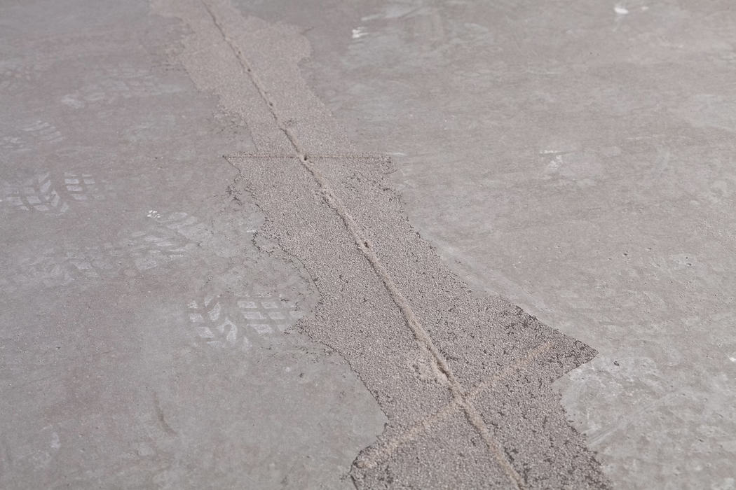 Nahaufnahme eines grauen Betonbodens mit einer diagonal verlaufenden, gefüllten Fuge, umgeben von grobem Reparaturmaterial und schwachen Schuhsohlen- oder Reifenspuren; sichtbare Struktur und kleine Partikel.