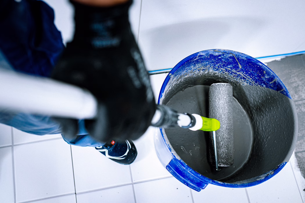 Draufsicht auf einen blauen Kunststoffeimer, der graue Farbe oder Beschichtung enthält, in dem eine Farbrolle liegt. Eine Person mit dunklem Handschuh und blauer Arbeitskleidung hält den Verlängerungsgriff der Rolle; ein weiß gefliester Boden und Arbeitsschuhe sind sichtbar. Der Eimerrand ist mit getrockneten Farbspritzern bedeckt, die Rollenhülse ist gleichmäßig mit grauem Material überzogen.