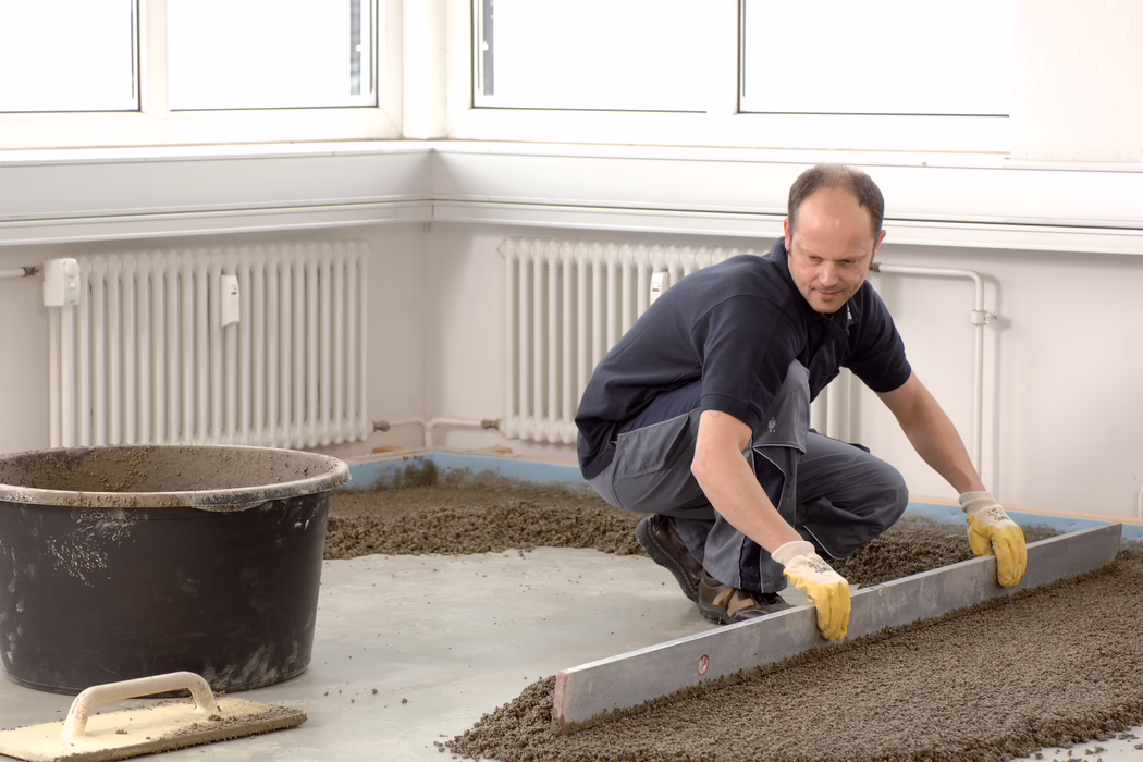 UZIN_Application_Image_Turbolight-System_2018-03 Ein Bauarbeiter in Arbeitskleidung kniet auf einem Betonboden in einem hellen Raum und nivelliert frisch aufgetragenen Estrich oder Zement mit einer langen Richtlatte. Er trägt gelbe Handschuhe und Arbeitsschuhe; in der Nähe stehen ein großer Mischkübel und eine Glättkelle. Weiße Heizkörper und Fenster an den Wänden deuten auf eine Innenrenovierung hin; der Arbeiter konzentriert sich darauf, eine gleichmäßige, glatte Fläche zu erzeugen.