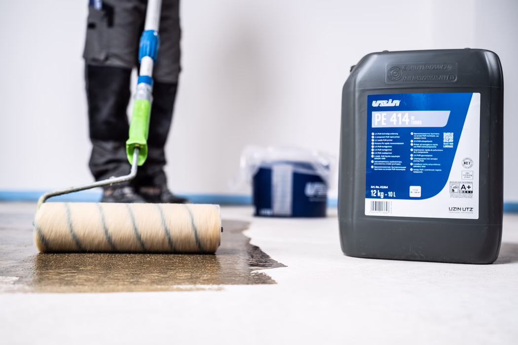 Nahaufnahme eines Handwerkers, der mit einer großen Farbwalze eine braune Bodenschicht auf einem hellen Betonboden aufträgt; ein großer grauer Kanister mit der Aufschrift 'PE 414' und ein blauer Eimer stehen daneben; die Beine und Werkzeuge des Arbeiters sind zu sehen, das Gesicht ist nicht sichtbar.