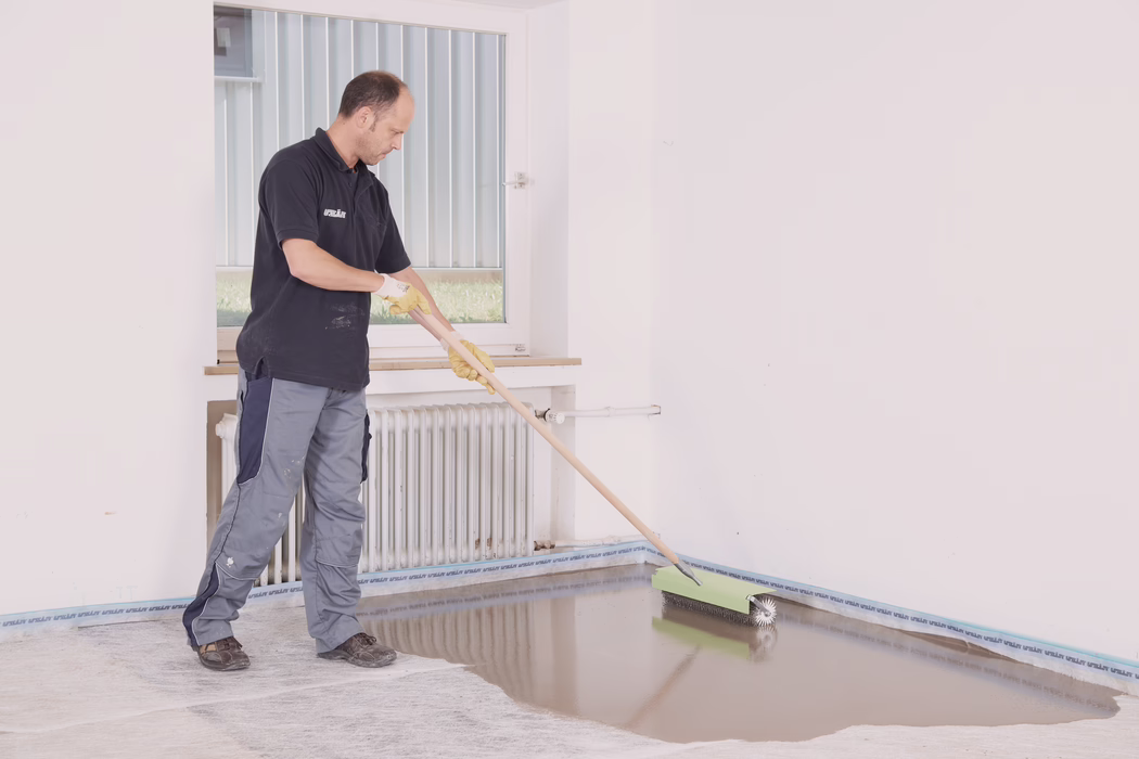 UZIN_Application_Image_Turbolight_MW02680_adapted_2018-10 Ein Handwerker in Arbeitshose, dunklem Polohemd und gelben Handschuhen verteilt mit einer langstieligen Stachelwalze eine selbstverlaufende Bodenspachtelmasse in einem leeren, hellen Raum. Er führt die Walze entlang der Fußbodenkante in der Nähe eines weißen Heizkörpers und eines Fensters und erzeugt eine glatte, glänzende, noch nasse Fläche, die Licht spiegelt. Die Wände sind weiß, an der Sockelleiste ist Schutzklebeband angebracht, und auf der Arbeitskleidung sind Farbflecken sichtbar; die Szene deutet auf eine professionelle Bodenverlegung oder Renovierung hin.