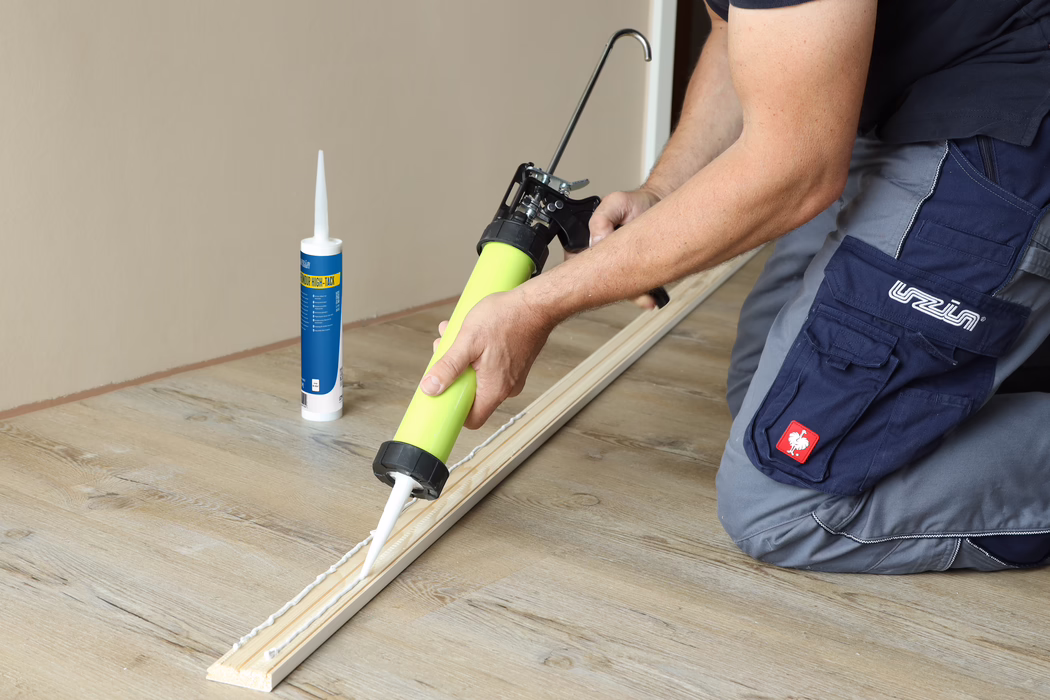 Nahaufnahme eines Handwerkers, der mit einer Kartuschenpistole weißen Baukleber auf eine Holzleiste auf einem hellen Holzboden aufträgt. Eine Kleberkartusche steht daneben. Der Handwerker kniet und trägt blaue Arbeitshosen; das Bild fokussiert Hände, Pistole und die Klebenaht.