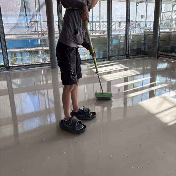 A person wearing shorts and sandals uses a roller tool to spread a coating on a shiny indoor floor, with sunlight streaming through large windows.