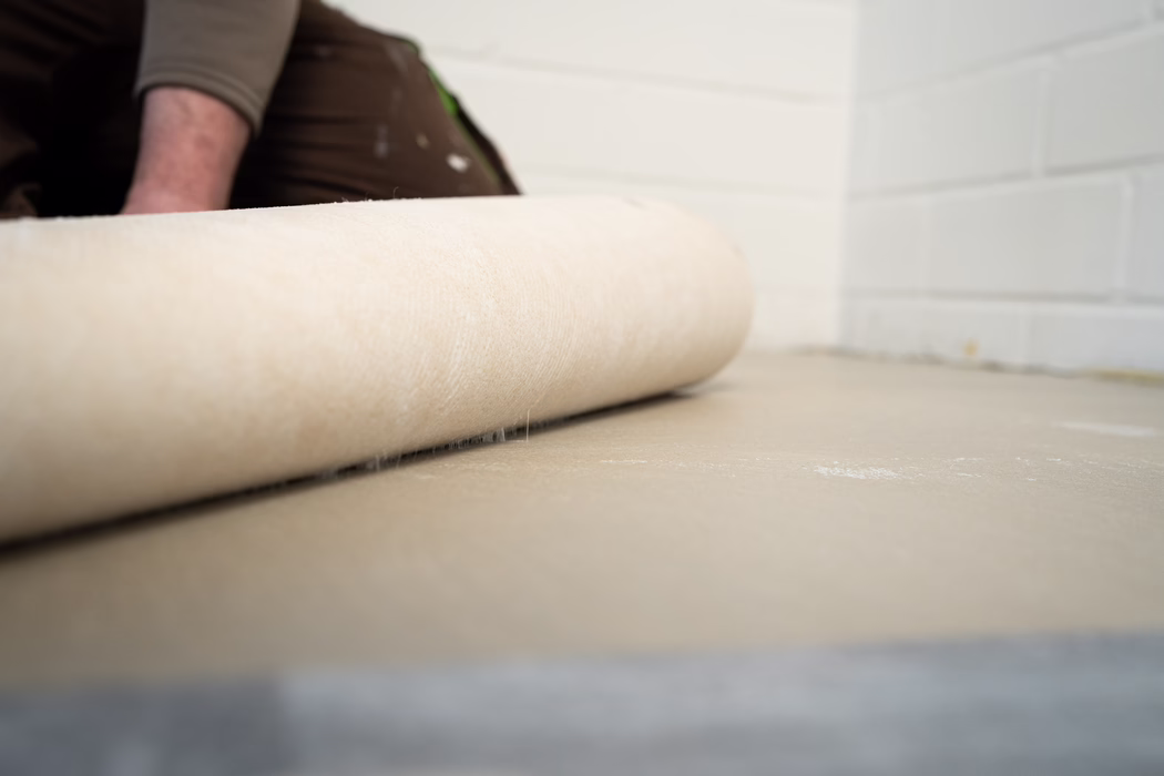 Nahaufnahme einer beigen Teppichunterlage, die auf einem Betonboden in einem Raum mit weiß gestrichenen Betonblockwänden ausgerollt wird; im Hintergrund ist teilweise der Arm eines Arbeiters und eine braune Arbeitshose zu sehen.