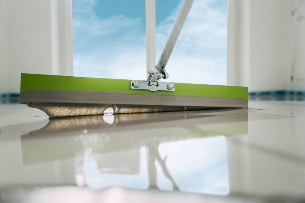 Nahaufnahme aus niedriger Perspektive eines Fußbodenspachtels mit grünem Rand, der glänzende selbstverlaufende Masse oder Epoxid auf einem hellen Boden glattzieht. Der Metallgriff und die Scharnierbefestigung sind im oberen Bereich sichtbar, zwei Schrauben halten die Halterung. Die feuchte, glatte Masse bildet eine dünne Kante und spiegelt das offene Fenster und den blauen Himmel, wodurch auf der frisch geglätteten Oberfläche eine klare spiegelähnliche Reflexion entsteht. Weiches Tageslicht betont feine Texturen und kleine Unebenheiten im Material; die geringe Schärfentiefe hält den Werkzeugkopf scharf, während Vorder- und Hintergrund sanft unscharf sind. Die Szene verweist auf eine Innenrenovierung oder Bodenfinish und hebt den Kontrast zwischen der grünen Kante des Werkzeugs, dem metallischen Beschlag und der cremigen, reflektierenden Bodenfläche hervor.
