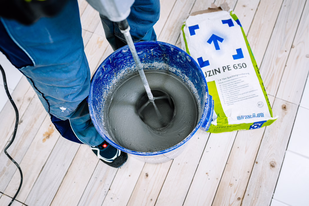 Aufsicht auf einen Handwerker, der eine graue Ausgleichsmasse in einem blauen Plastikeimer mit einem Rührgerät anmischt. Der Arbeiter trägt blaue Arbeitshosen und schwarze Sneakers und steht auf hellen Holzdielen. Eine neben dem Eimer liegende Tüte mit der Aufschrift 'UZIN PE 650' ist zu sehen, während ein Mixaufsatz eines Elektromixers die glatte graue Paste rührt. Die Szene wirkt wie bei Renovierungsarbeiten: Staub, ein Stromkabel auf dem Boden und am rechten Bildrand sind weiße Fliesen erkennbar.