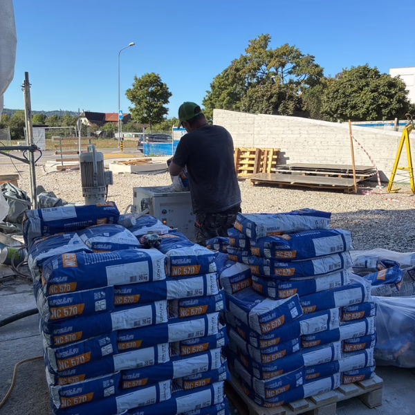 A worker stands near stacked bags of construction material at an outdoor construction site with machinery and trees in the background under a clear blue sky.