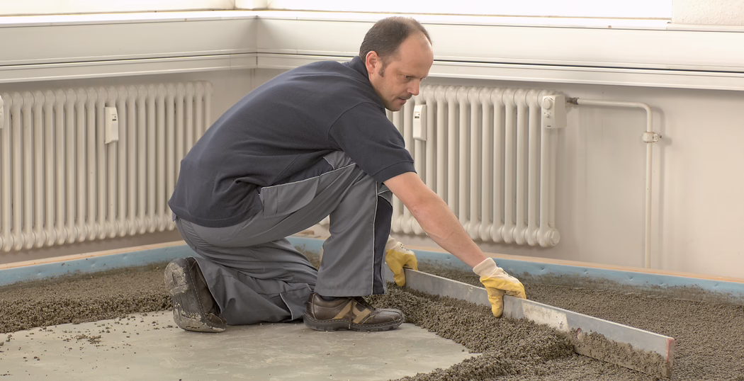 UZIN_Application_Image_SC 914 Turbo_2018-03 A construction worker kneels on a concrete floor inside a bright room, wearing a navy shirt, gray work trousers and yellow gloves. He is using a long metal straightedge to level freshly laid cement or floor screed along the wall beneath a white radiator. The scene shows wet aggregate and tools on the floor, protective edge insulation at the perimeter, and natural light coming from a high window. The worker is focused on flattening and smoothing the surface to prepare it for the next flooring layer.